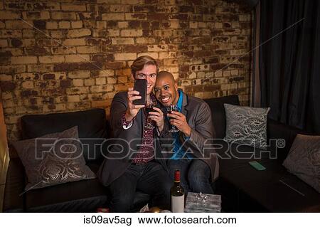 Stock Image - Male couple sitting on sofa, taking self portrait using smartphone. Fotosearch