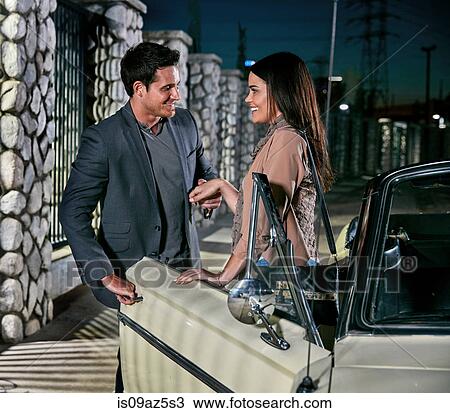 Stock Image - Man helping woman out of classic car at night, Los Angeles, California, USA. Fotosearch