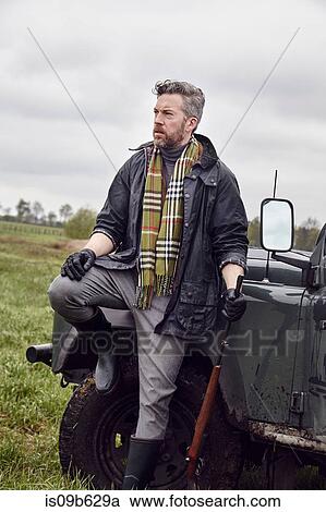 Stock Image - Man hunting, leaning against off road vehicle in field with shot gun. Fotosearch