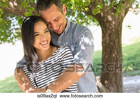 Stock Photograph - Man in front of tree with arms around woman smiling. Fotosearch