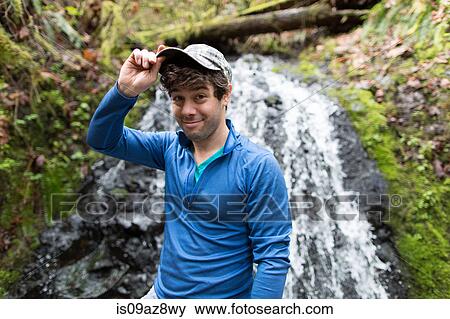 Stock Photo - Man in front of waterfall wearing baseball cap looking at camera smiling. Fotosearch