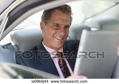 Picture - Mature businessman in back of car, using digital tablet. Fotosearch