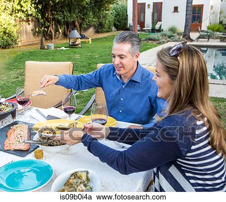 Stock Image - Mature couple serving food at garden table. Fotosearch