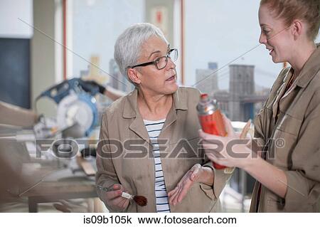 Stock Image - Mature female carpenter explaining wood stain to trainee in furniture making workshop. Fotosearch
