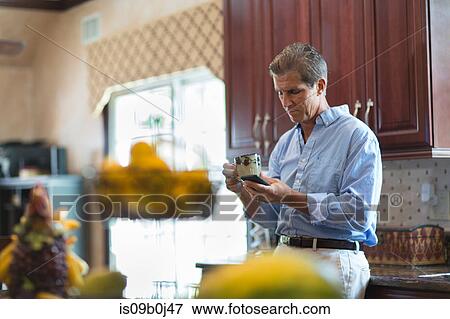 Mature man in kitchen, holding coffee, using smartphone View Large Photo Image Stock Photo - Mature man in kitchen, holding coffee, using smartphone. Fotosearch