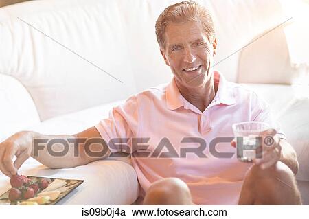 Stock Image - Mature man relaxing at home, holding drink, eating strawberries. Fotosearch