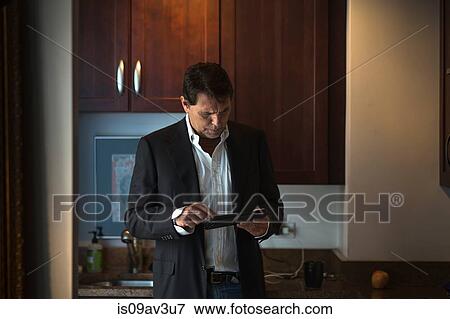 Mature man wearing suit jacket in kitchen, holding digital tablet looking down View Large Photo Image Stock Photo - Mature man wearing suit jacket in kitchen, holding digital tablet looking down. Fotosearch