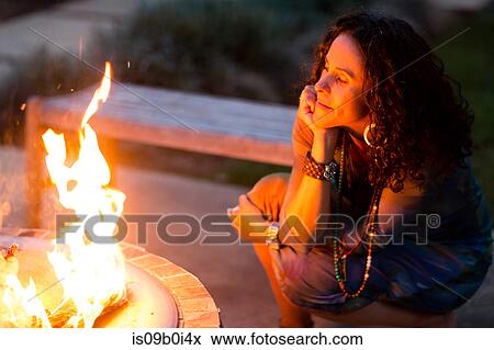 Stock Photo - Mature woman crouching and watching patio fire at dusk. Fotosearch