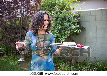 Stock Image - Mature woman holding cutting board and red wine at garden party. Fotosearch