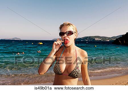 Stock Image - Mature woman in leopard print bikini licking ice lolly on beach, Ibiza, Spain. Fotosearch