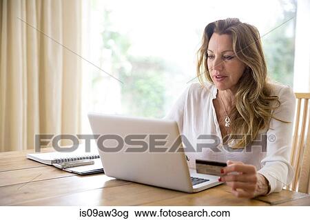 Stock Image - Mature woman using credit card with laptop at home. Fotosearch