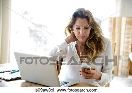 Mature woman using laptop whilst reading smartphone text at home View Large Photo Image Stock Image - Mature woman using laptop whilst reading smartphone text at home. Fotosearch