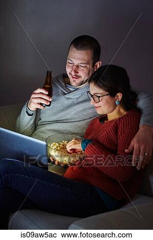 Mid adult couple sitting on sofa in evening eating popcorn and looking at laptop View Large Photo Image Stock Image - Mid adult couple sitting on sofa in evening eating popcorn and looking at laptop. Fotosearch