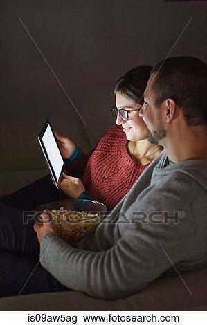 Mid adult couple sitting on sofa in evening eating popcorn and looking at digital tablet View Large Photo Image Stock Image - Mid adult couple sitting on sofa in evening eating popcorn and looking at digital tablet. Fotosearch