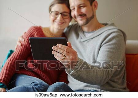 Stock Photography - Mid adult couple sitting on sofa taking selfie with digital tablet. Fotosearch