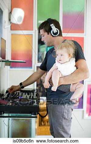 Picture - Mid adult man cooking, listening to headphones and carrying baby daughter. Fotosearch