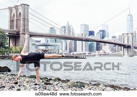 Stock Image - Mid adult man practicing yoga on riverside in front of Brooklyn Bridge, New York, USA. Fotosearch