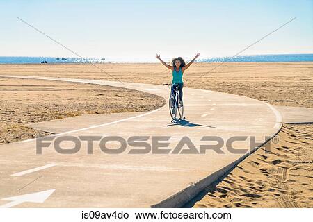 Mid adult woman cycling along pathway at beach, arms in air View Large Photo Image Picture - Mid adult woman cycling along pathway at beach, arms in air. Fotosearch