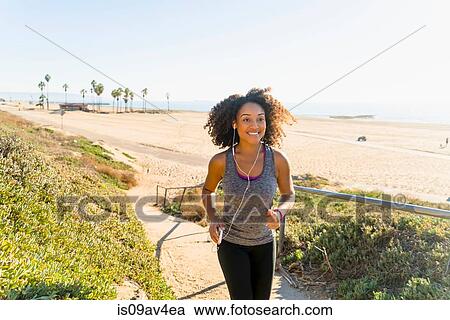 Stock Image - Mid adult woman running along pathway by beach. Fotosearch