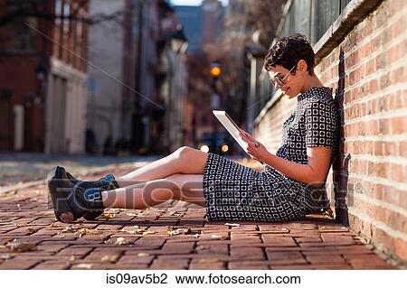 Stock Image - Mid adult woman sitting against brick wall, using digital tablet. Fotosearch
