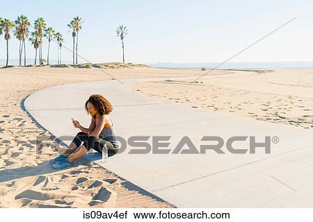 Mid adult woman sitting by beach, using smartphone View Large Photo Image Stock Image - Mid adult woman sitting by beach, using smartphone. Fotosearch
