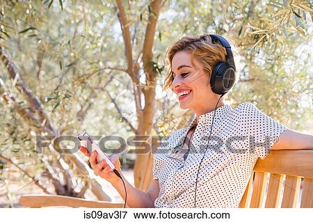 Mid adult woman sitting on bench, wearing headphones, looking at smartphone, smiling View Large Photo Image Stock Photo - Mid adult woman sitting on bench, wearing headphones, looking at smartphone, smiling. Fotosearch