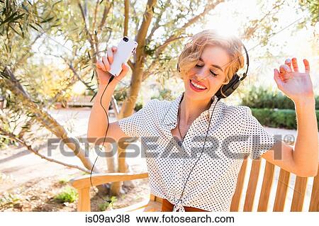 Stock Photo - Mid adult woman sitting on bench, wearing headphones, holding smartphone, dancing. Fotosearch