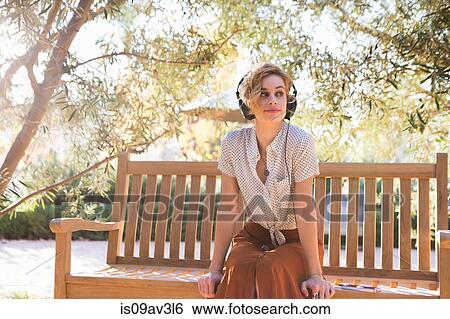 Stock Photograph - Mid adult woman sitting on bench, wearing headphones, looking away. Fotosearch