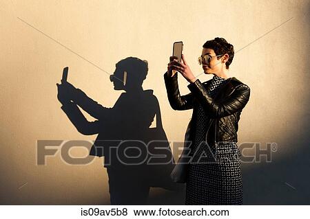 Mid adult woman taking self portrait, using smartphone, shadow cast on wall beside her View Large Photo Image Stock Photo - Mid adult woman taking self portrait, using smartphone, shadow cast on wall beside her. Fotosearch