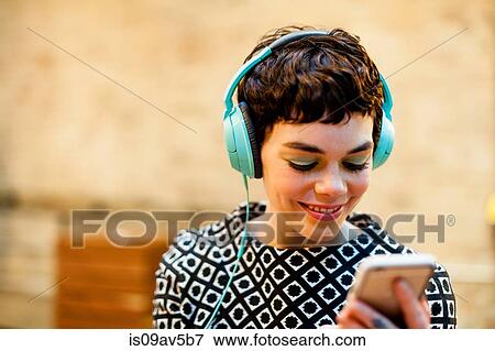 Stock Photo - Mid adult woman, wearing headphones, looking at smartphone, smiling. Fotosearch