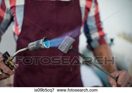 Stock Photo - Mid section of man heating up branding iron with blow torch in factory. Fotosearch