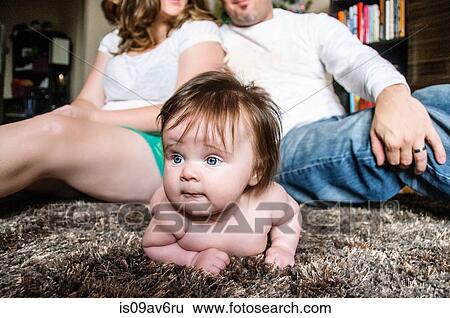 Stock Photograph - Mother and father sitting back to back on rug with baby girl lying on rug looking away. Fotosearch