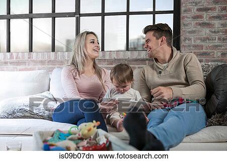Mother and father sitting on sofa with toddler, toddler holding digital tablet View Large Photo Image Picture - Mother and father sitting on sofa with toddler, toddler holding digital tablet. Fotosearch