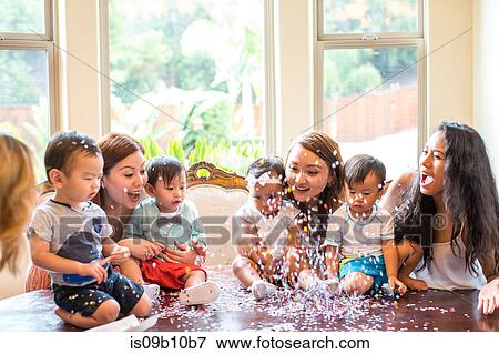Stock Photo - Mothers with baby girl and boys celebrating with confetti on dining room table. Fotosearch