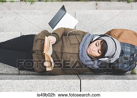 Stock Image - Overhead view of female backpacker lying on back reading book on wall. Fotosearch