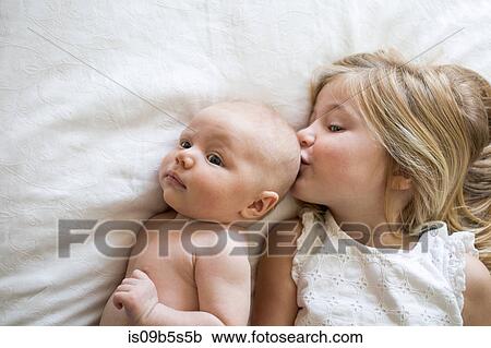 Stock Image - Overhead view of young girl and baby brother lying on bed, girl kissing baby brother. Fotosearch