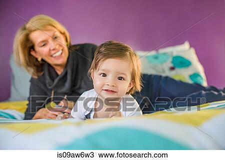 Stock Image - Portrait of baby boy in front of grandmother reclining on bed. Fotosearch