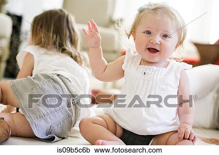 Stock Image - Portrait of baby girl sitting on floor with sister behind. Fotosearch