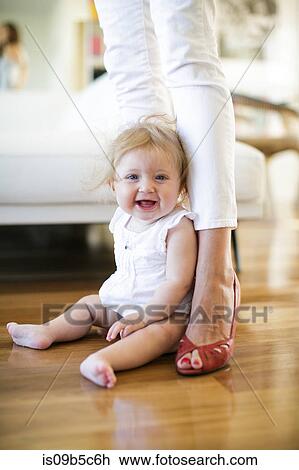 Stock Image - Portrait of baby girl sitting on floor between mother's legs. Fotosearch