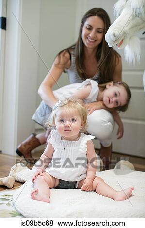 Stock Photo - Portrait of baby girl sitting on floor with mother and sister behind. Fotosearch