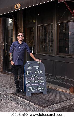 Stock Image - Portrait of barman leaning against happy hour board outside public house window, Brooklyn, New York, USA. Fotosearch