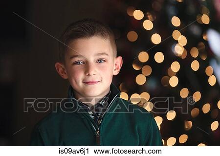 Stock Image - Portrait of boy in front of christmas tree looking at camera smiling. Fotosearch