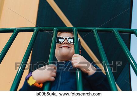 Stock Image - Portrait of boy wearing sunglasses looking through railings smiling. Fotosearch
