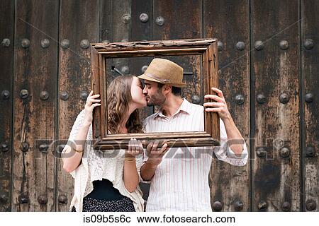 Stock Image - Portrait of couple, kissing, holding wooden frame in front of their faces, Mexico City, Mexico. Fotosearch