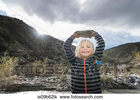 Stock Image - Portrait of cute boy holding up rock from riverbed. Fotosearch