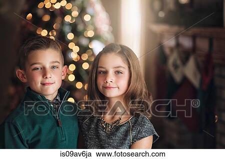 Stock Image - Portrait of girl and boy in front of christmas tree looking at camera smiling. Fotosearch