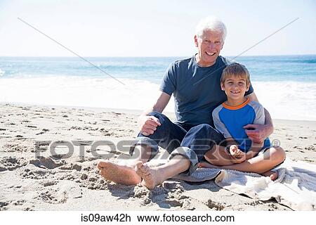 Stock Image - Portrait of grandfather and grandson, sitting on beach, smiling. Fotosearch