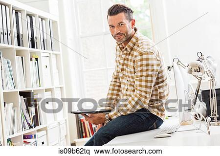 Stock Photography - Portrait of male designer sitting on desk in creative studio. Fotosearch