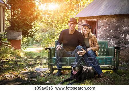Stock Photograph - Portrait of mature couple with dog, sitting on bench, outdoors. Fotosearch