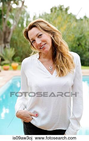 Stock Image - Portrait of mature woman with long blond hair with hand in pocket in front of swimming pool. Fotosearch